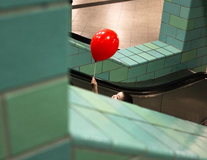 Child on escalator holding a red balloon in teal tiled urban setting, capturing street photos of unexpected moments.