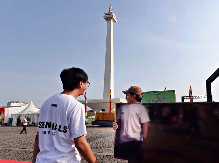 Two men in a city square with a tall monument, captured in a perfectly timed street shot showing a reflection.