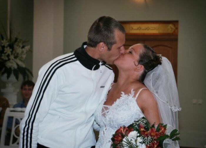 Bride in a wedding dress and veil kissing groom in a white track jacket, creating an awkward wedding photo moment.