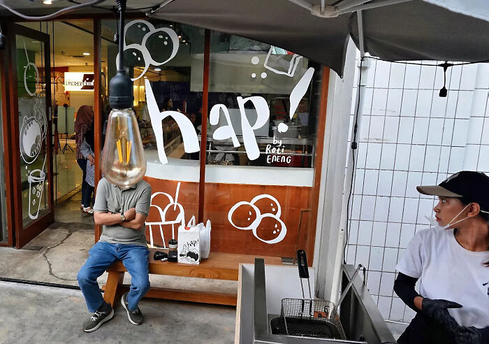 Man sitting with arms crossed by cafe window, a hanging light bulb perfectly timed in this street photography shot.