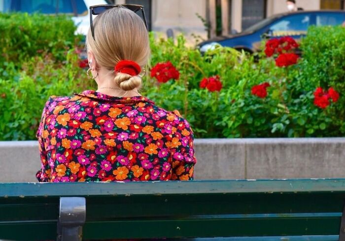Street photographer captures a woman in a colorful floral shirt sitting on a bench with red flowers in background.