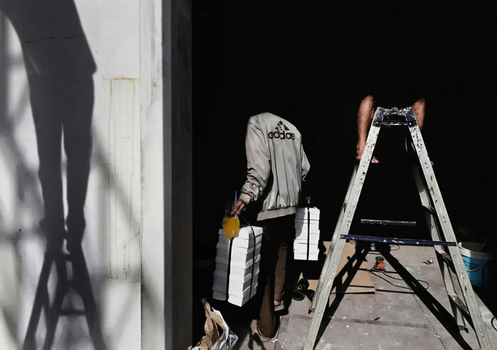 Photographer captures perfectly timed street shot of person carrying stacked boxes with ladder and shadow nearby at work site