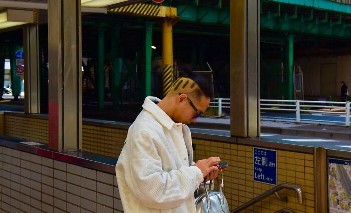 Street photographer captures perfect coincidences of a man with unique hair design checking his phone at an urban train station.