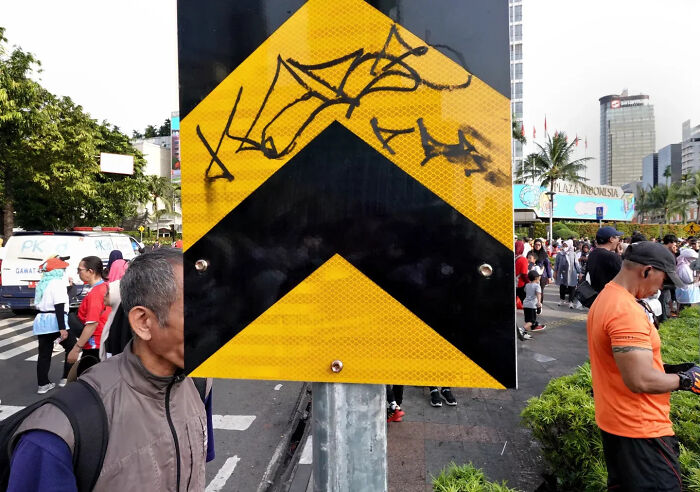 Street shot perfectly timed with a man walking behind a graffiti-covered yellow and black traffic sign in a busy urban area.