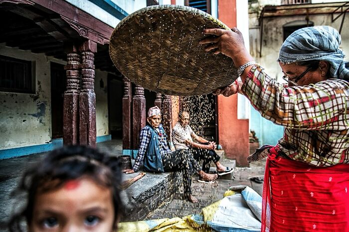Candid street photo showing a woman sifting grains with others sitting in a rustic setting, capturing humanity and tender moments.