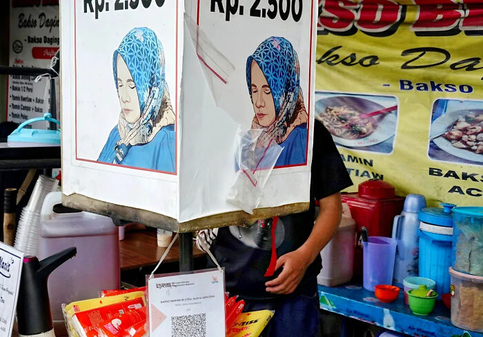 Street shot of a food vendor behind a sign with a woman in a blue headscarf, capturing perfectly timed street photography moments.