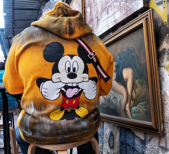 Man wearing a vibrant Mickey Mouse hoodie sits beside a framed painting in a candid street photo capturing humanity and absurdity.