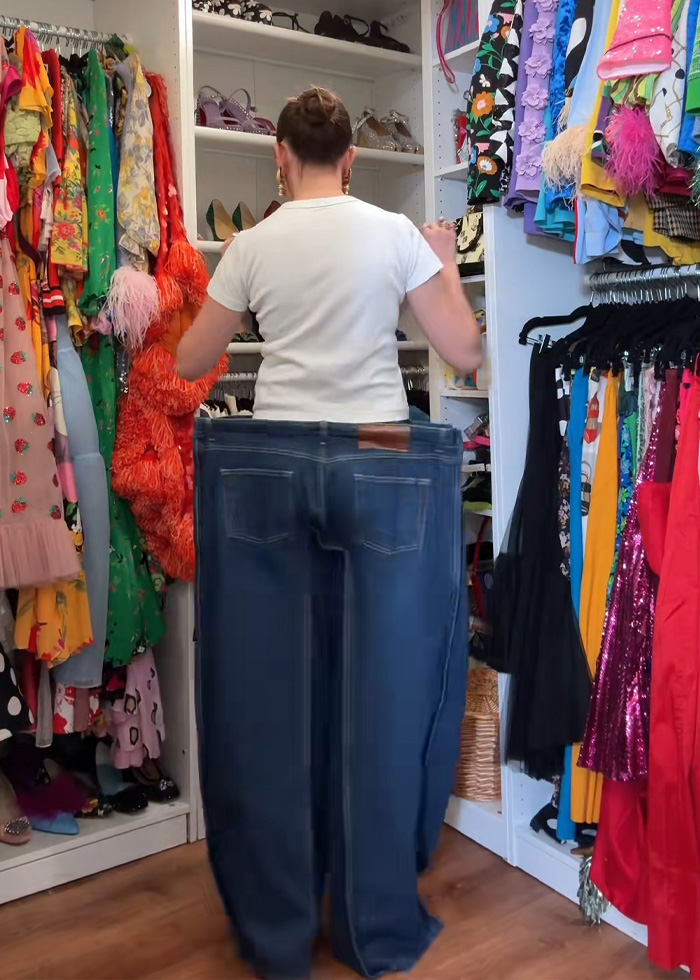 Woman in white shirt wearing oversized cubic jeans inside a colorful walk-in closet with various dresses hanging around.