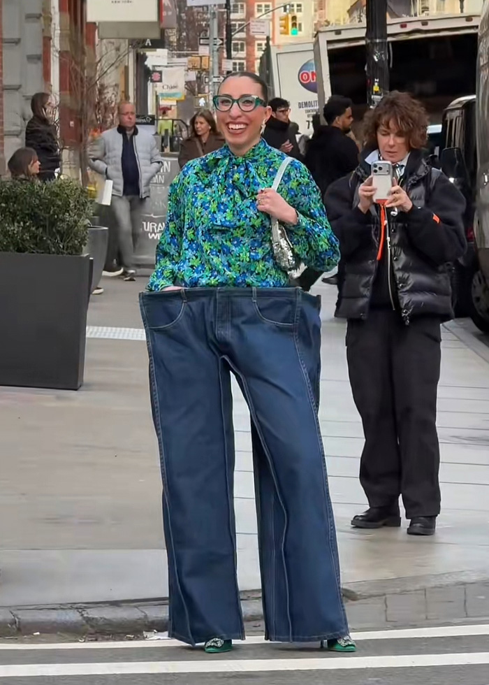 Influencer wearing oversized cubic jeans and a floral blouse, smiling on a city street while being photographed.