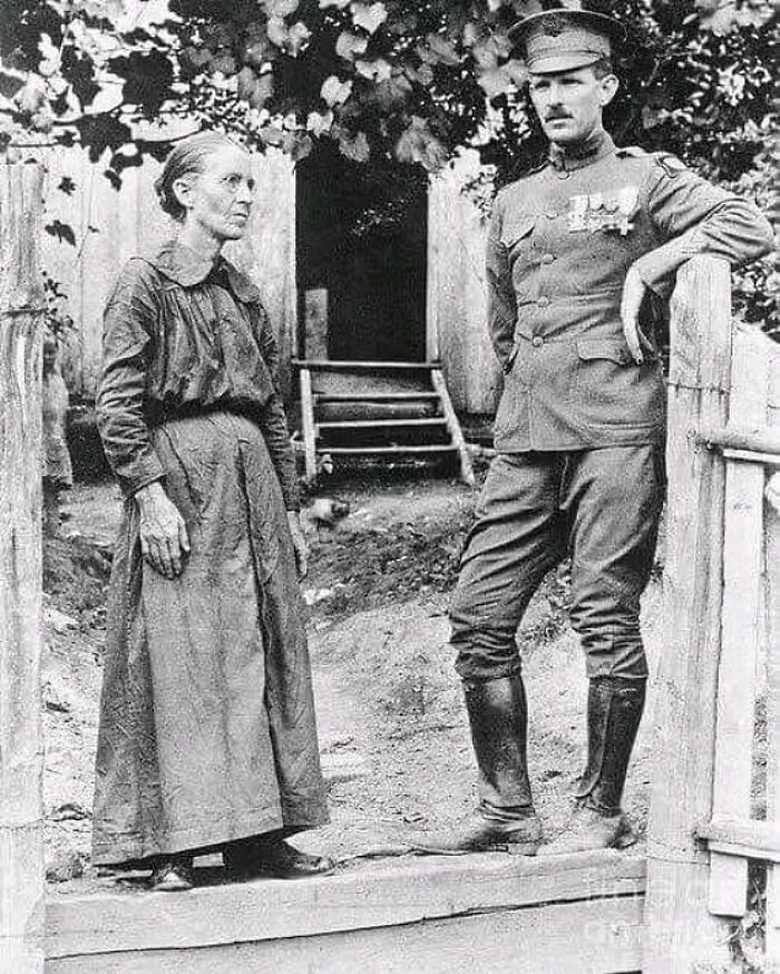 Black and white historical photograph of a soldier in uniform standing by a wooden gate with an elderly woman, depicting humanity's sad history.