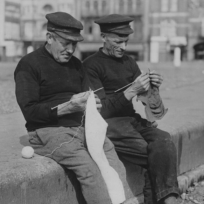 Two men knitting outdoors in a historic black and white photograph showing the sad, beautiful history of humanity.