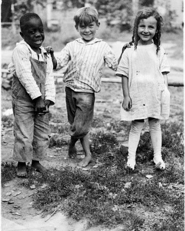 Three children from diverse backgrounds smiling and playing barefoot in a historic photograph showcasing humanity's past.