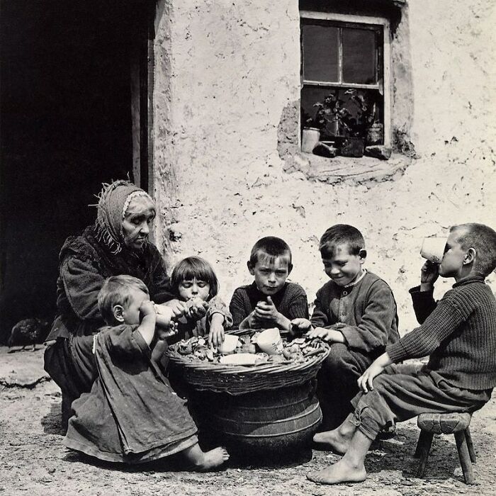 Black and white photo of an elderly woman and children sharing a simple meal, reflecting the sad history of humanity.