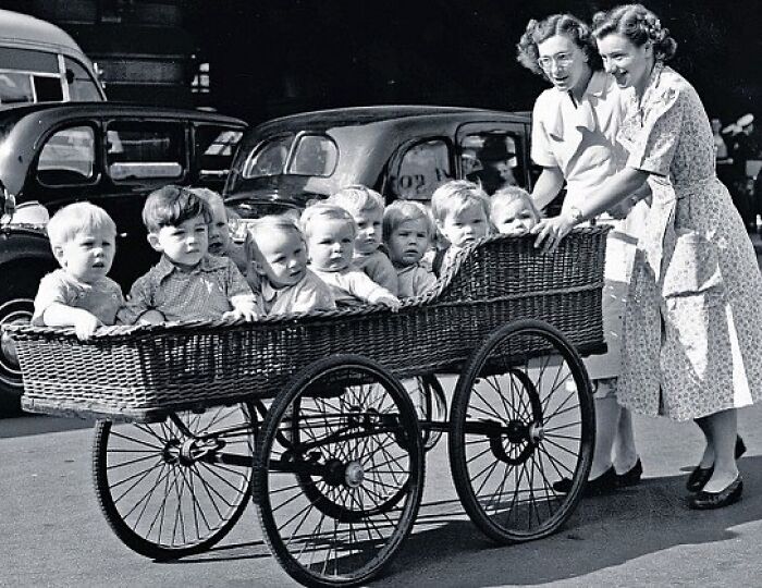 Black and white historical photograph showing women pushing a large vintage stroller with multiple children, reflecting humanity history.