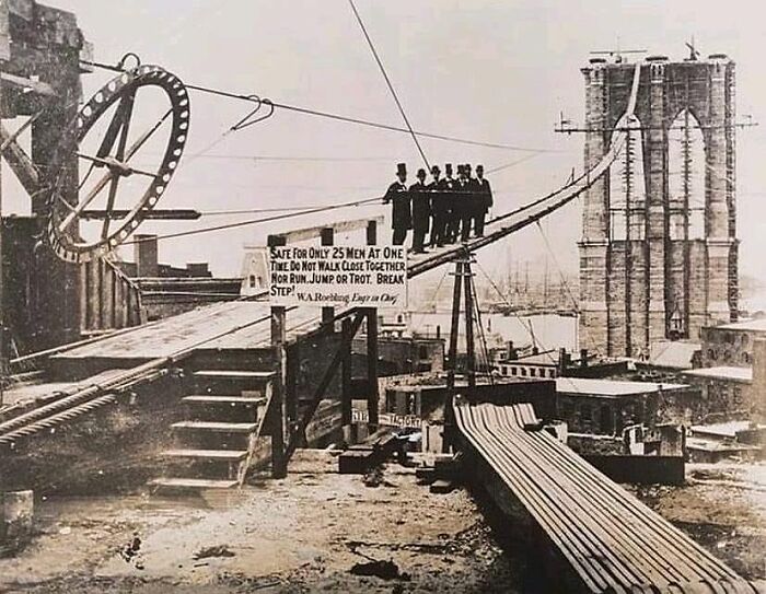Historic photograph showing workers on a suspension bridge cable illustrating the complex history of humanity and labor risks.