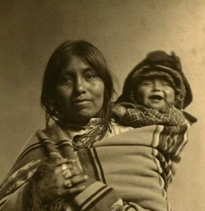 Native woman carrying a baby wrapped in traditional blanket, a powerful photograph showing history of humanity.