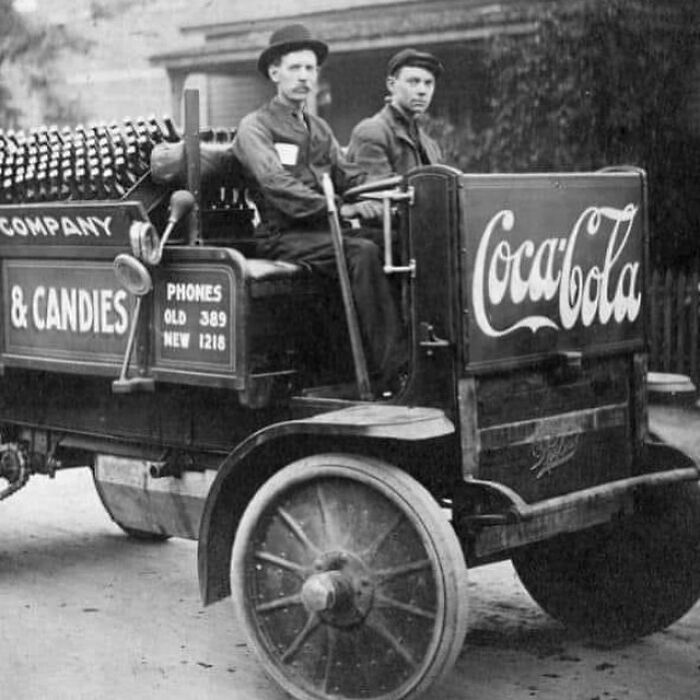 Two men riding an early 20th-century Coca-Cola delivery truck, capturing a historic moment in the history of humanity.