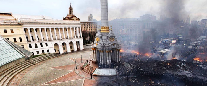 Monument and city square showing a dramatic before and after scene with fire and smoke on one side and intact buildings on the other.