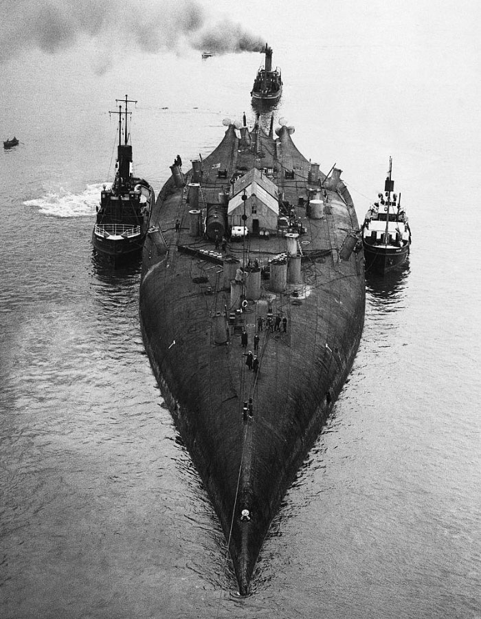 Historic black and white photo of a large submarine escorted by boats, showing a side of history often missing from textbooks.