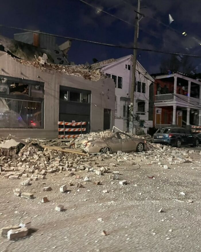 Damaged car covered in bricks and rubble on a street with collapsed building debris in front of houses at night.