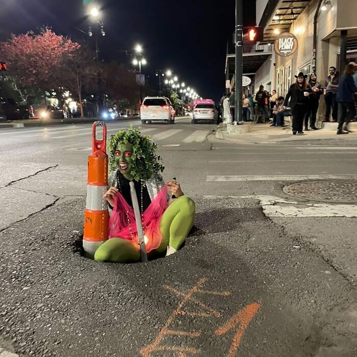 Person dressed as a plant sitting in a street pothole next to a traffic cone, one of the weirdest things found in the streets.