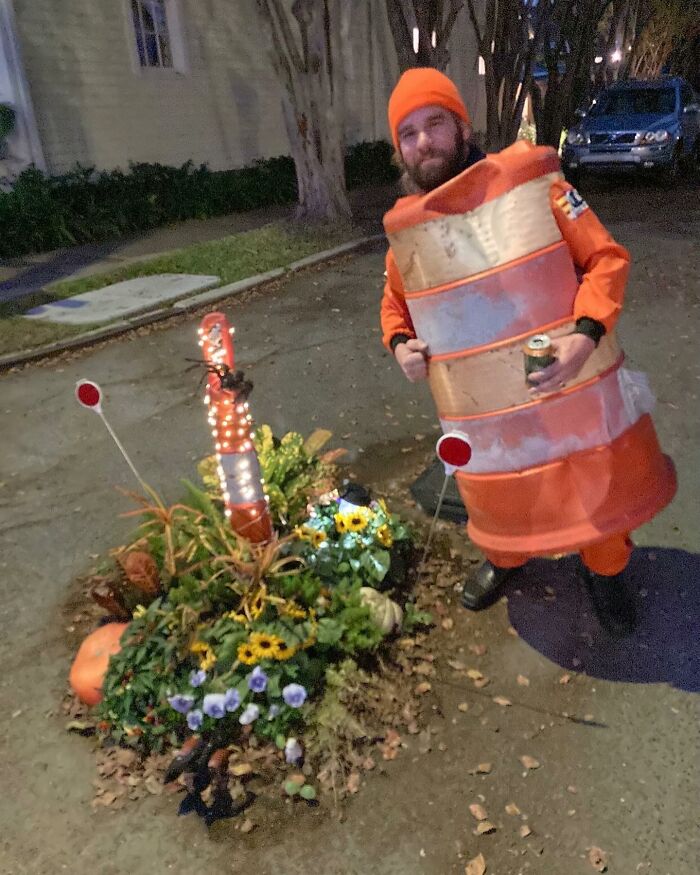 Man dressed as an orange traffic cone standing next to decorated traffic cone on street with flowers at night