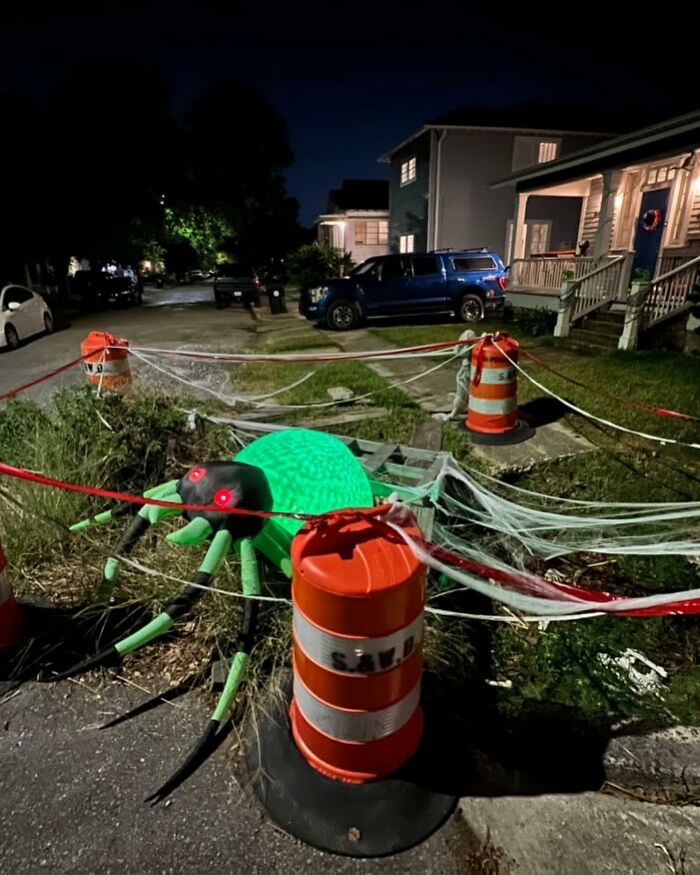 Giant glowing green spider with red eyes surrounded by orange cones and caution tape found in the street at night.