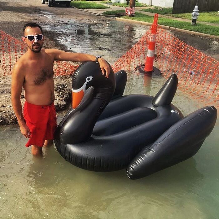 Man wearing sunglasses and red shorts standing in flooded street next to a large inflatable black swan, a weird street find.