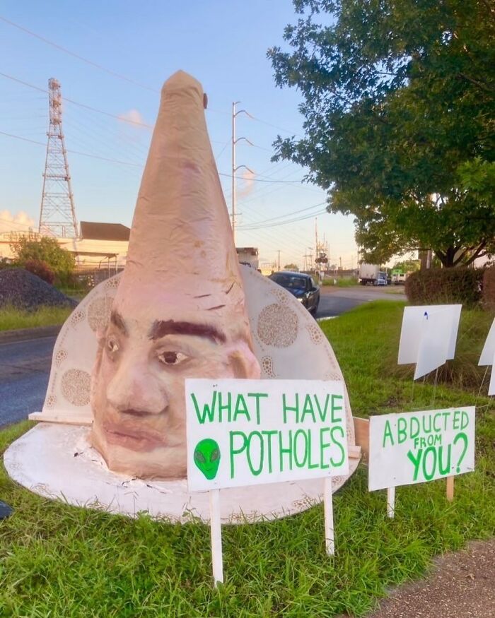 Large cone-shaped sculpture with a human face near street signs asking about potholes and abduction, weirdest things found.