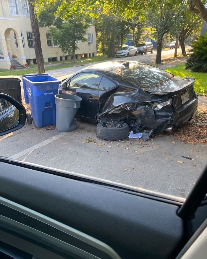 Damaged black car with flat tire parked oddly next to trash bins on a residential street, showcasing weirdest things found in streets.