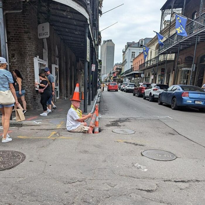 Man wearing a traffic cone hat sitting partially in a manhole on a city street, a weird street find among pedestrians.