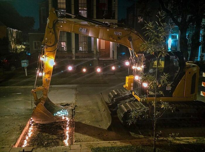 Yellow excavator decorated with string lights digging a rectangular hole on a residential street at night, unusual street find.