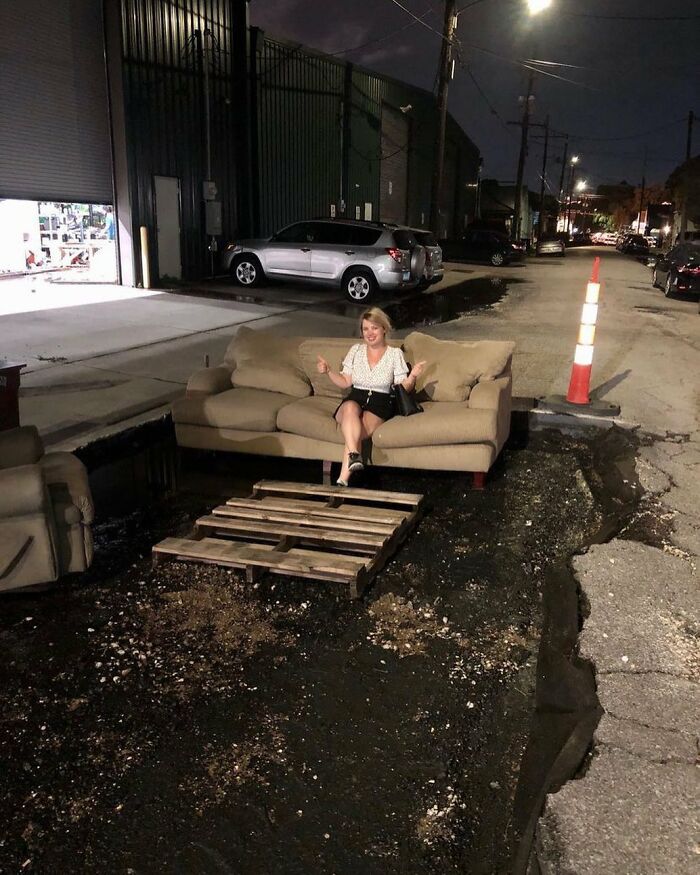 Woman sitting on a couch placed in a street pothole with a wooden pallet bridge, showcasing weird street finds at night.