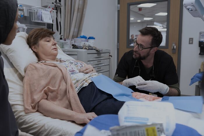 Hospital scene with a woman lying in bed and a doctor in gloves discussing the tragic true story in The Pitt Season 2.