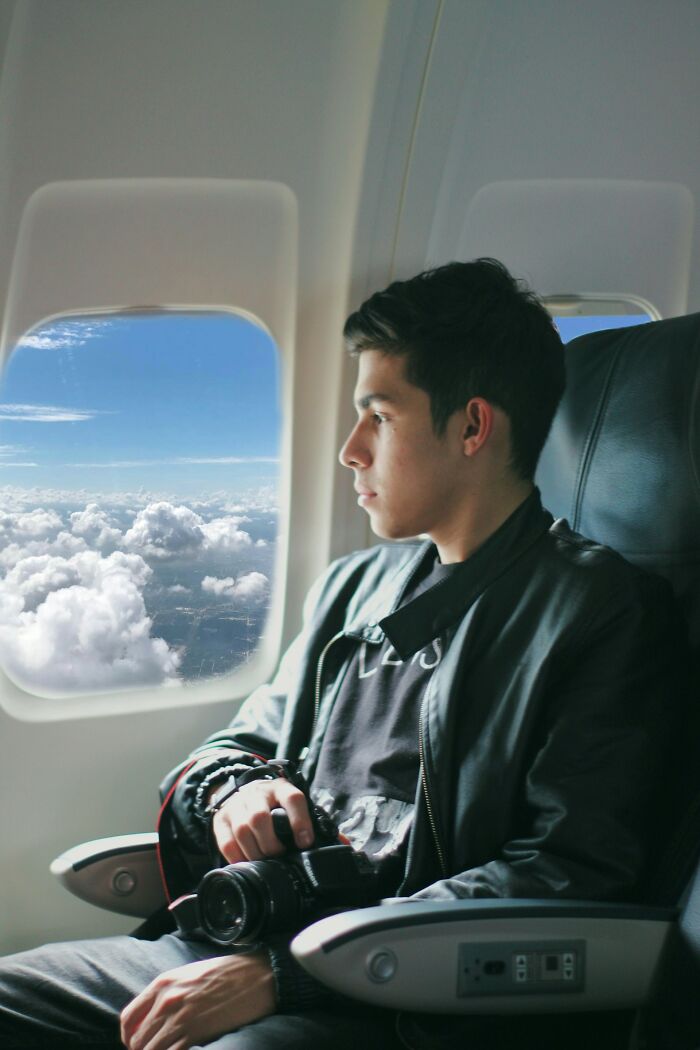 Young man holding a camera on a plane, looking out the window at clouds, highlighting rich people far from reality.