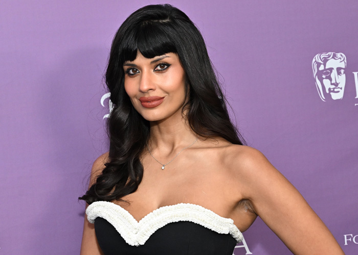 British actress Jameela Jamil posing at an event, wearing a black and white strapless dress against a purple backdrop.