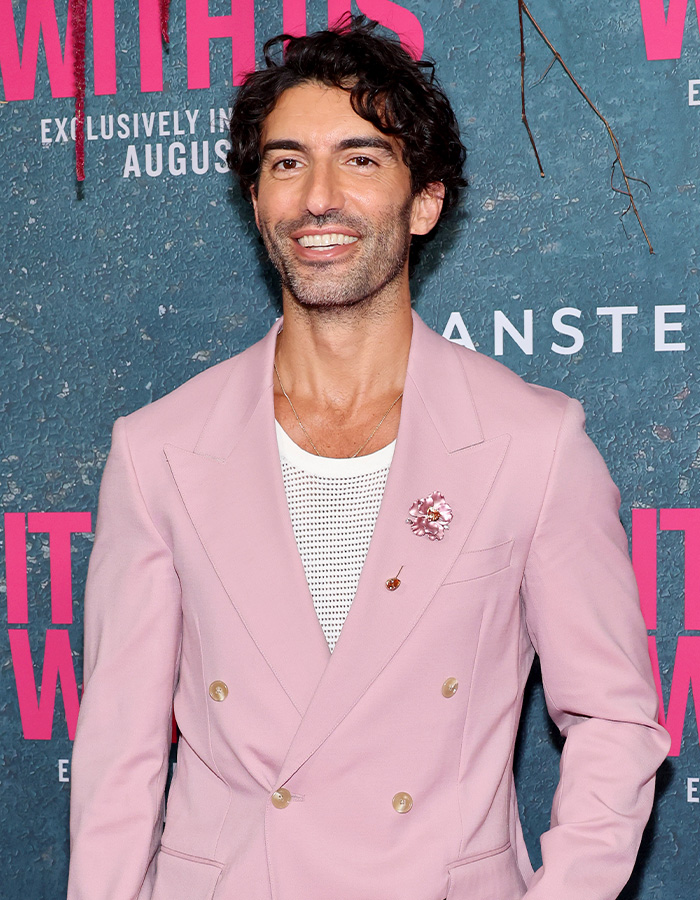 Man in a pink suit smiling at a media event with promotional backdrop featuring British actress Jameela Jamil and Blake Lively keywords.