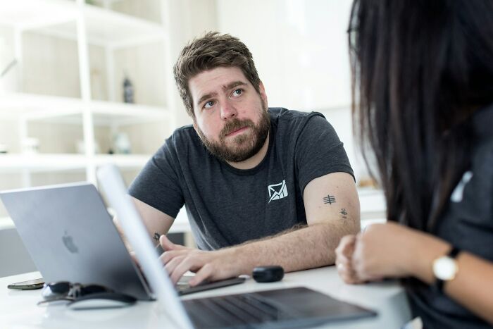 Man with tattoo listening intently during a discussion about former cult members and their experiences at a laptop-filled workspace.