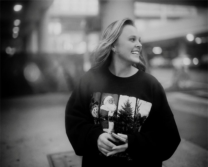 JoJo Siwa smiling outdoors in a casual sweatshirt, showcasing her dramatic transformation and name change recently.