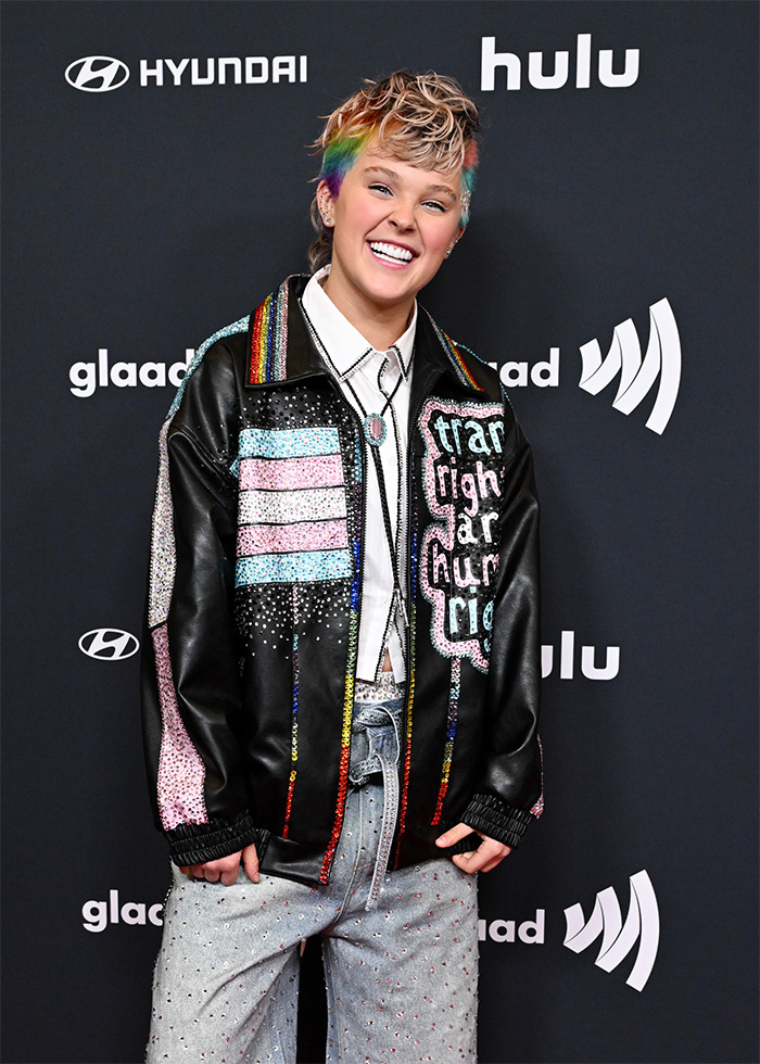 JoJo Siwa with rainbow hair wearing a colorful studded jacket at a red carpet event after dramatic transformation.