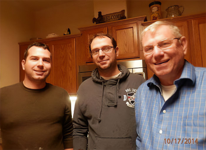 Three men standing in a kitchen, with the father of ICE agent who slew Renee Nicole Good smiling in a blue shirt.