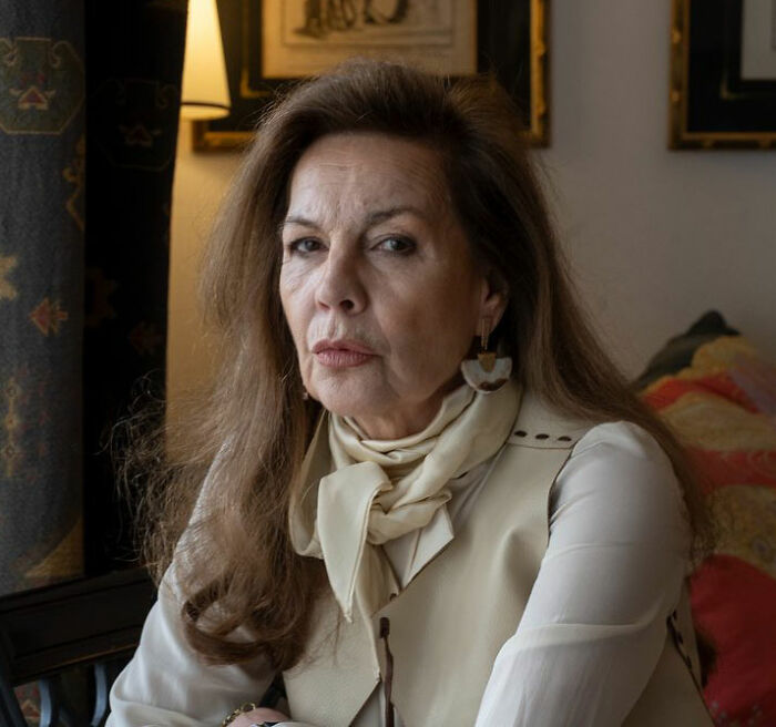 Older woman with long brown hair and serious expression seated indoors near a lamp and decorated wall.