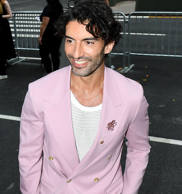 Justin Baldoni smiling in a light pink blazer with a white mesh shirt at an outdoor event with metal barriers behind him Justin Baldoni smiling in a light pink blazer with a white mesh shirt at an outdoor event with metal barriers behind him