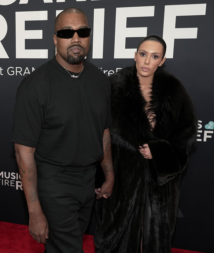 Kanye West wearing black sunglasses and outfit, posing with a woman in a black fur coat at an event.