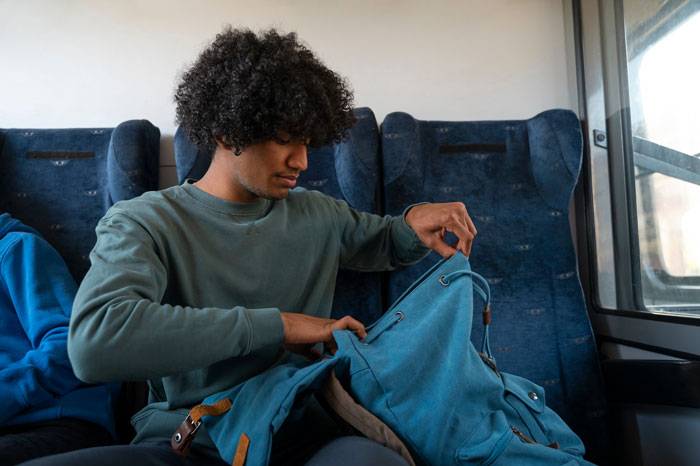 Teen on a crowded metro bus rummaging through his blue backpack while sitting near a window seat.