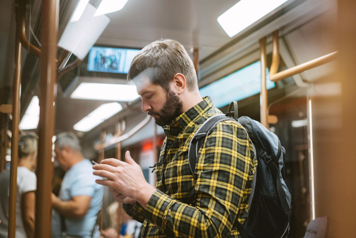 Young man with backpack standing in crowded metro train, focused on his phone, surrounded by other passengers.