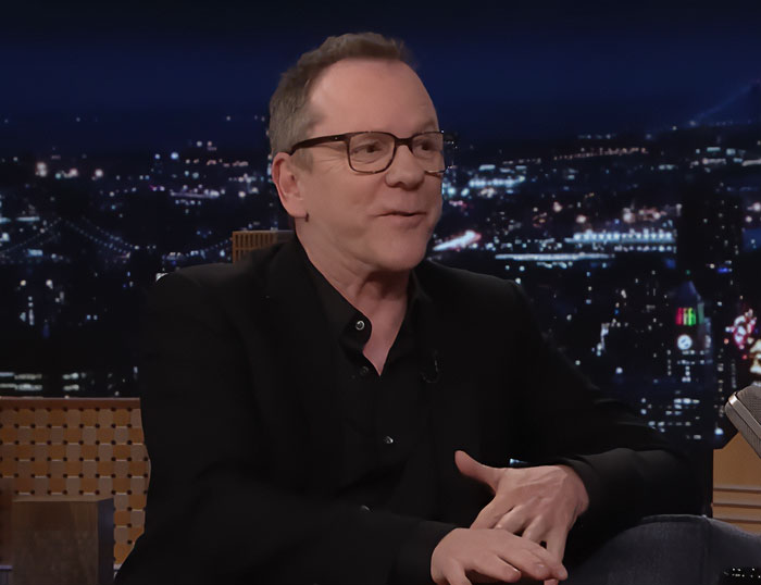 Kiefer Sutherland wearing glasses and a black jacket, speaking during a nighttime studio interview with city lights behind.