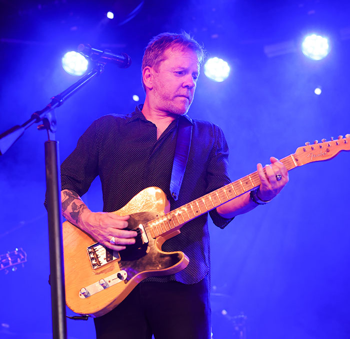 Kiefer Sutherland playing electric guitar on stage with blue lighting during a live music performance in Hollywood.
