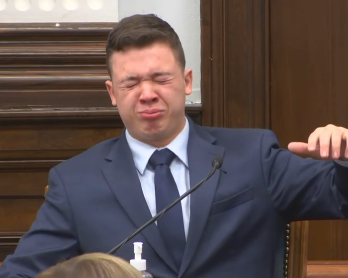 Kyle Rittenhouse in a courtroom wearing a suit, showing a pained expression during testimony about ICE slaying reaction.