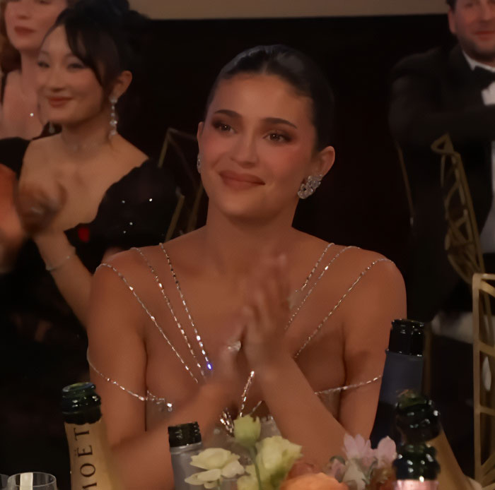 Kylie Jenner at the Golden Globes, dressed elegantly and clapping at an event with champagne bottles on the table.